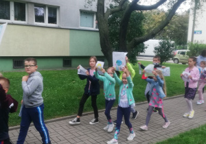 Zdjęcie przedstawia dzieci idące po chodniku z kolorowymi rysunkami w uniesionych rękach. Dzieci idą w luźnej grupie , pierwsza para trzyma transparent z napisem SPRZATANIE ŚWIATA. W tle widać okna bloku.